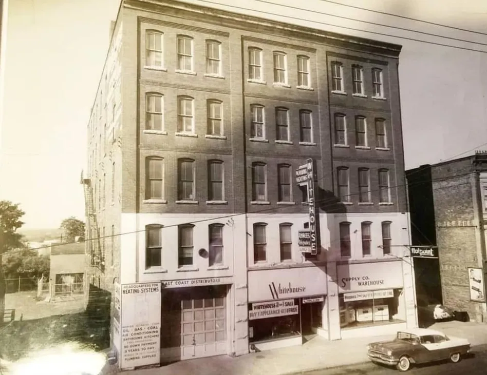 The building in the mid-1900s as the Whitehouse Supply Co., its storefronts painted with signs for heating systems and appliances, and a 1950s car parked out front