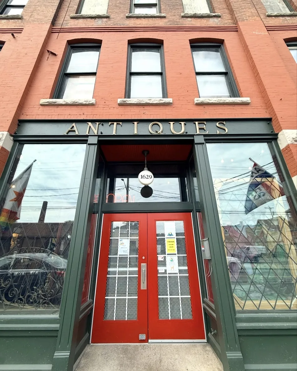 The Syracuse Antiques Exchange storefront entrance, with red double doors under brass ANTIQUES signage, a lit 1629 globe lamp overhead, and the brick upper floors rising above